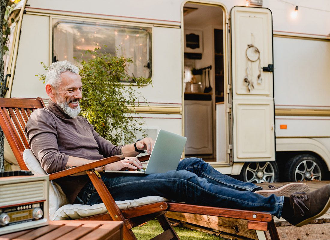 About Our Agency - Grey-Haired Man Resting on a Wooden Chair Using a Laptop With Caravan Van in the Background